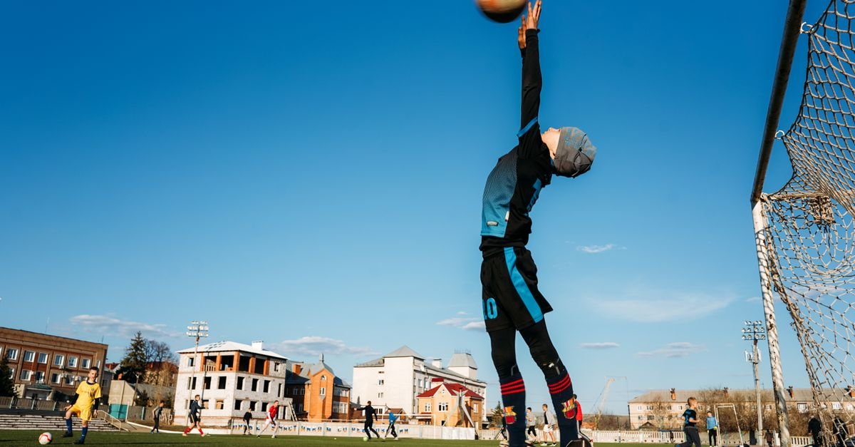 A child soccer player leaps to block a goal during an outdoor match on a sunny day.