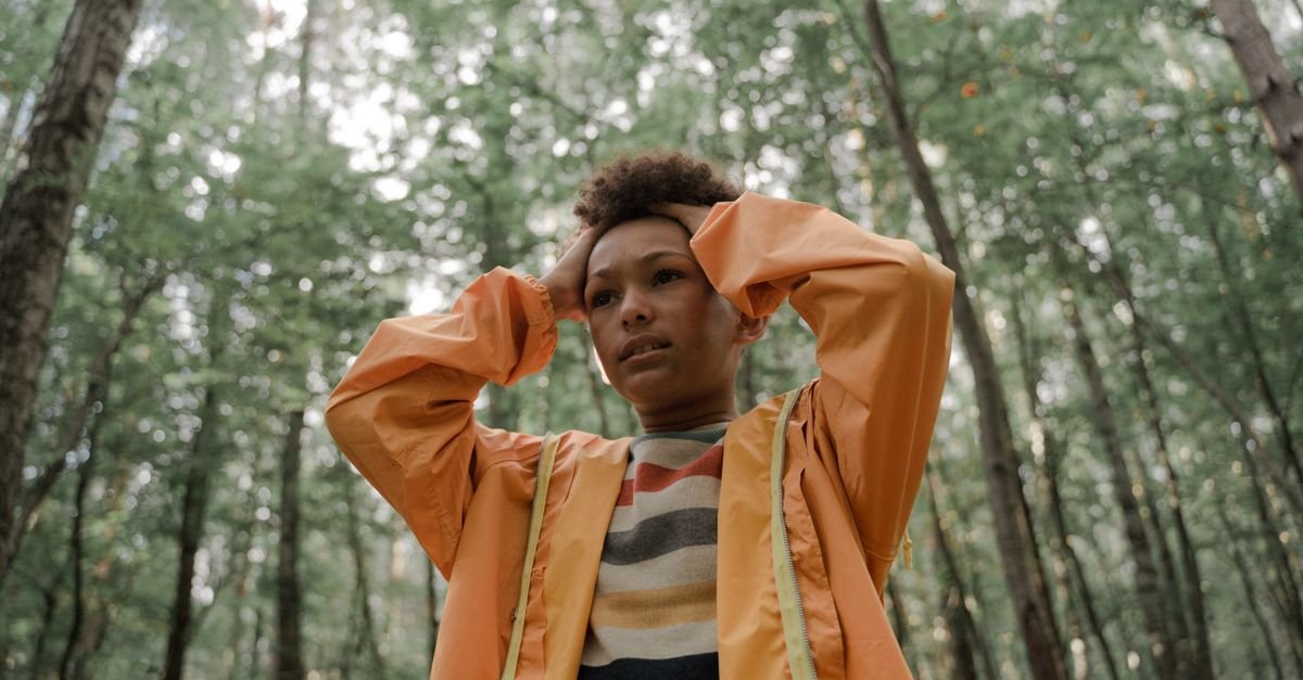 Teen holding head in thinking pose, wearing yellow raincoat, surrounded by forest greenery.