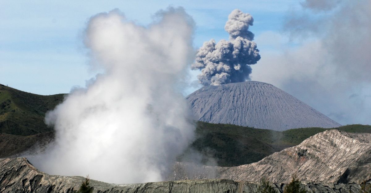 A dramatic aerial view of an erupting volcano with thick smoke plumes and rugged landscape.