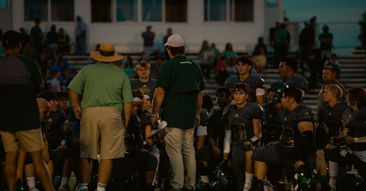 Football team huddles with coaches during a game at a stadium under the evening sky.