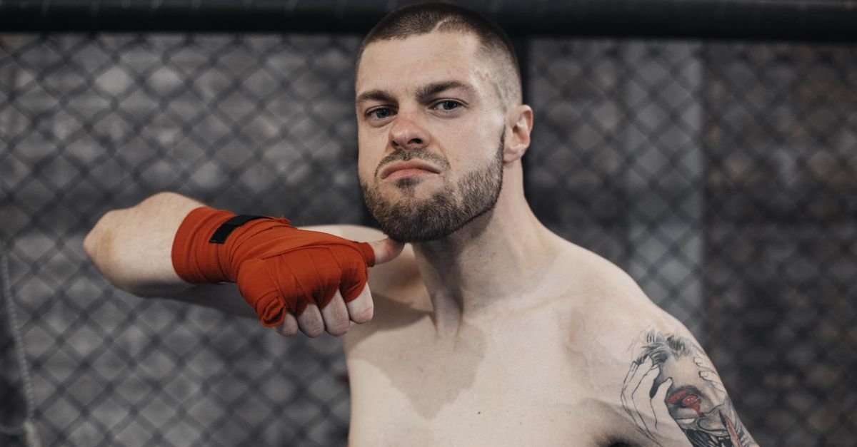 A muscular shirtless boxer with tattoos performing a chin flick gesture inside a cage.