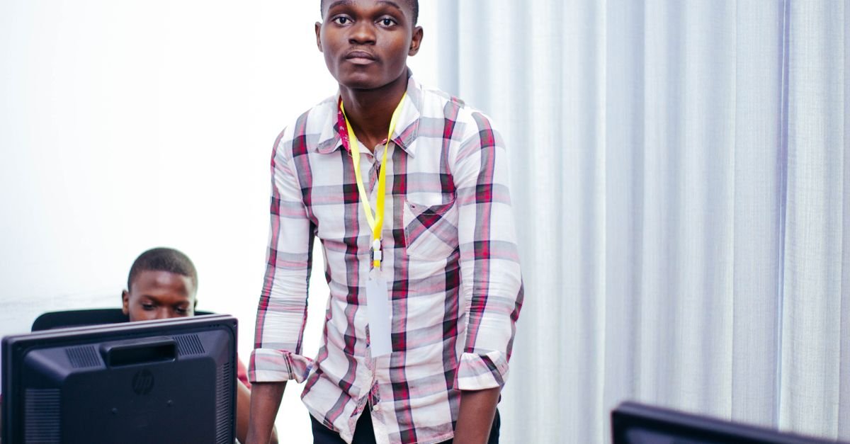 Young man in checked shirt engages in teaching session with computers in classroom.