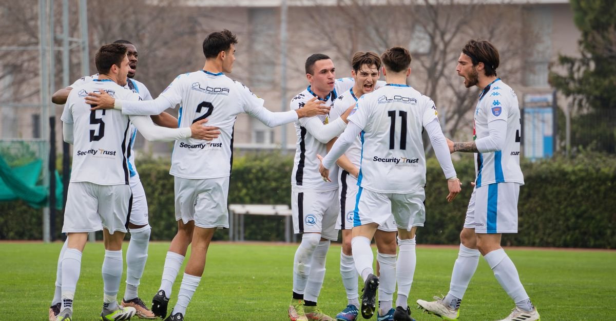 A group of soccer players in white uniforms celebrating on a soccer field during a daytime match.