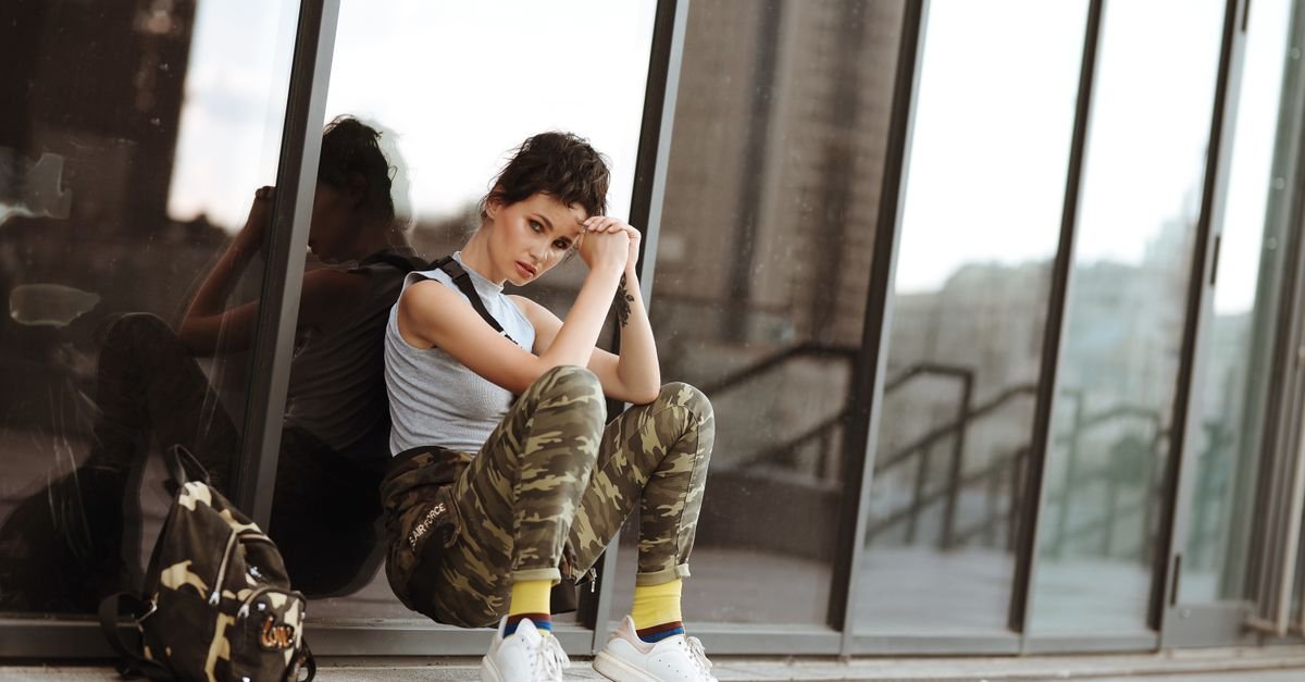 Young woman in urban setting wearing camo pants and white sneakers, leaning against a glass building.