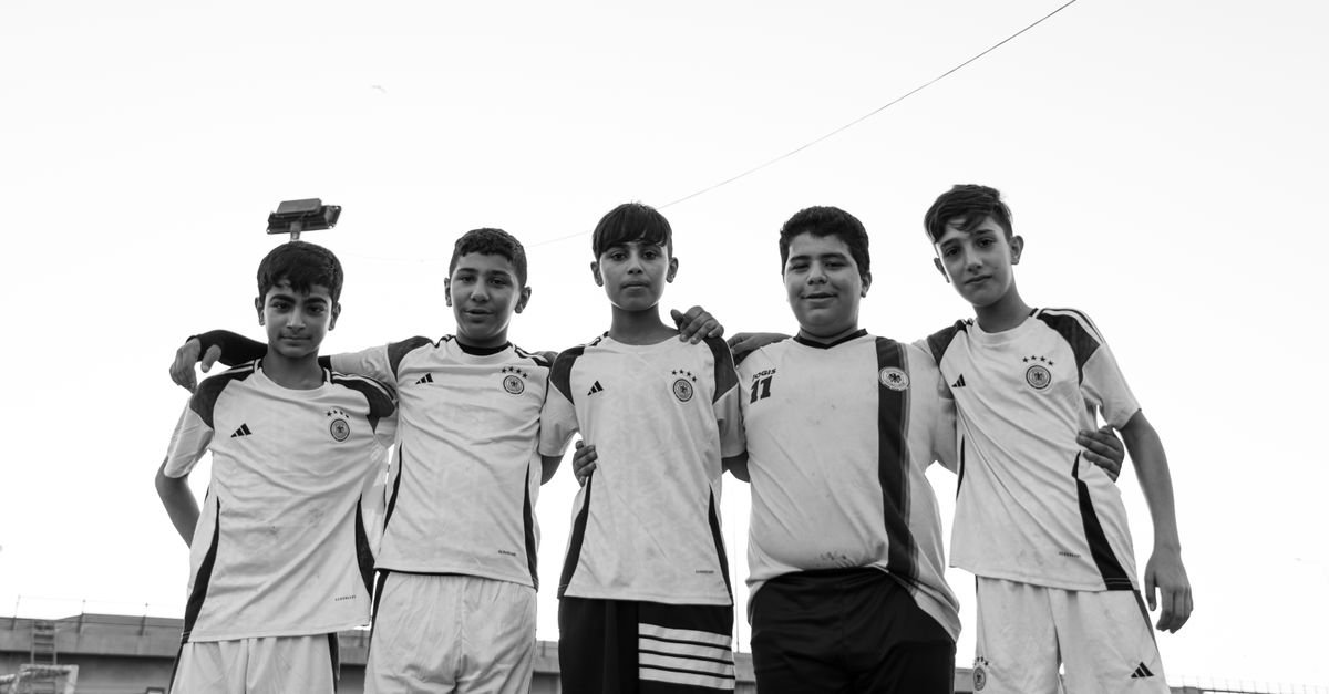 Five teenage boys in soccer jerseys smiling and embracing outdoors.