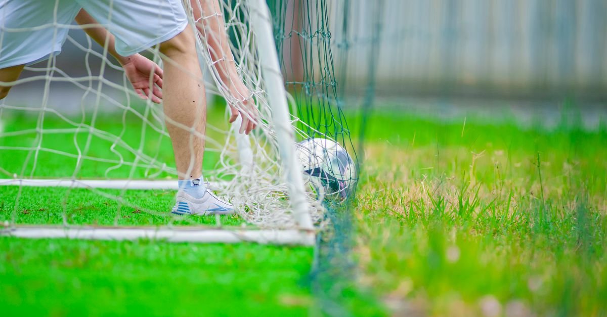 A soccer player retrieves a ball from the goal on a lush green field, captured in Hanoi, Vietnam.