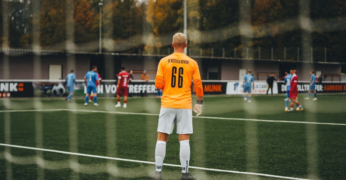 Goalkeeper in orange jersey stands ready during a soccer match on a fall day.