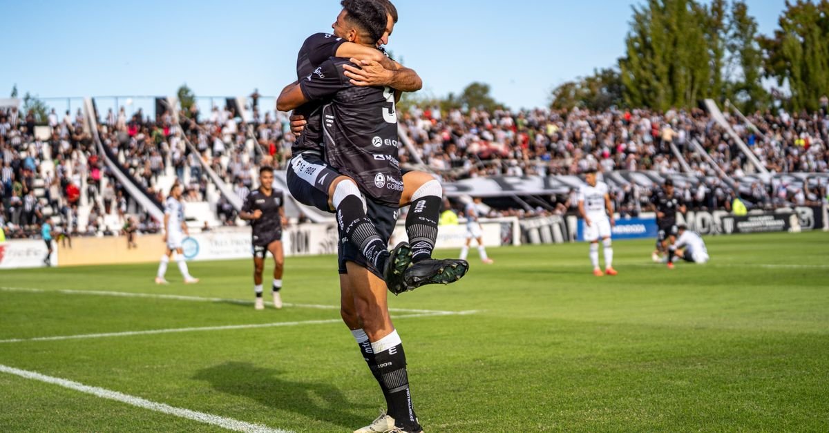 Two soccer players joyfully celebrate a goal during a match on a sunny day.