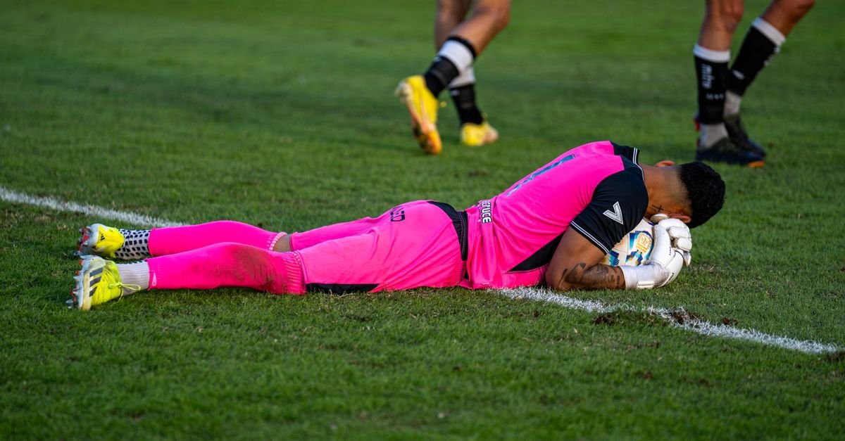 Soccer goalie in bright pink kit lying on grass field during a match.