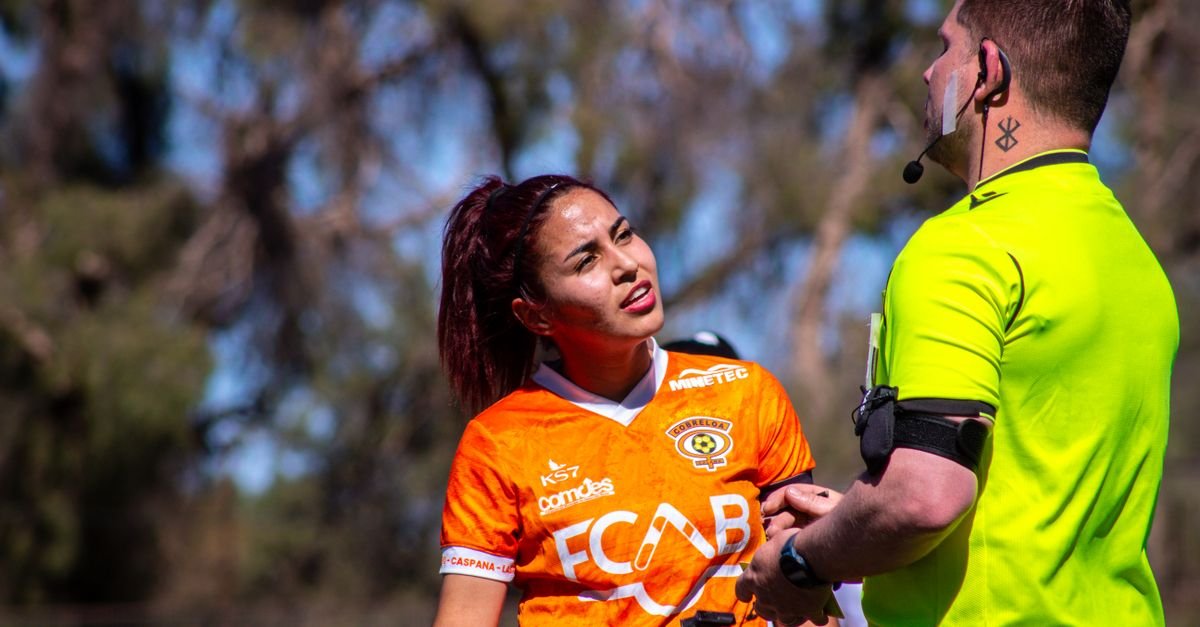 Female soccer player in orange jersey argues with referee on the outdoor field during a sunny day match.