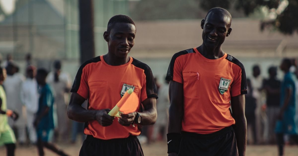 Two soccer referees in orange jerseys walking during a daytime match.
