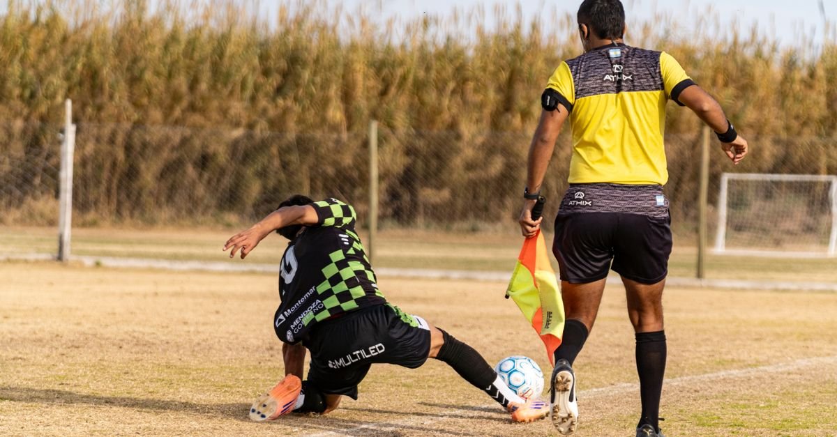 Soccer player tackles during a match as referee observes. Captured outdoors on a field.