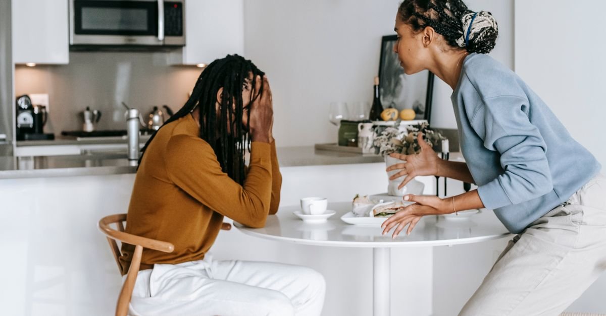 Young black man sitting at table with hands on head and having conflict with standing near table woman in light kitchen