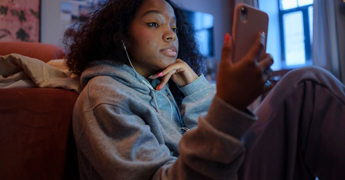 Teenage girl using smartphone indoors with earphones, wearing a hoodie, and looking relaxed.