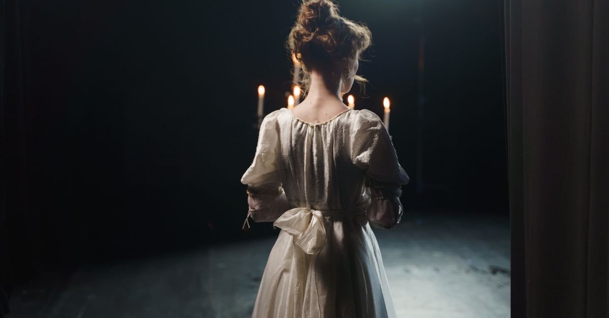 An actress in a white dress stands backstage in dim lighting, enhancing a classic theatrical atmosphere.
