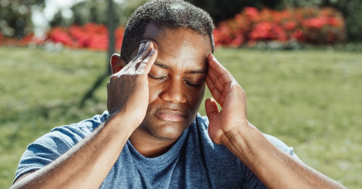 An adult man with eyes closed touches his temples, indicating a headache while seated outdoors.