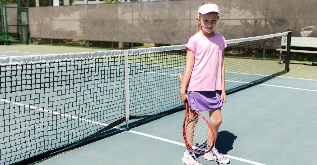 A girl in sportswear smiling on a tennis court holding a racket, enjoying a sunny day.