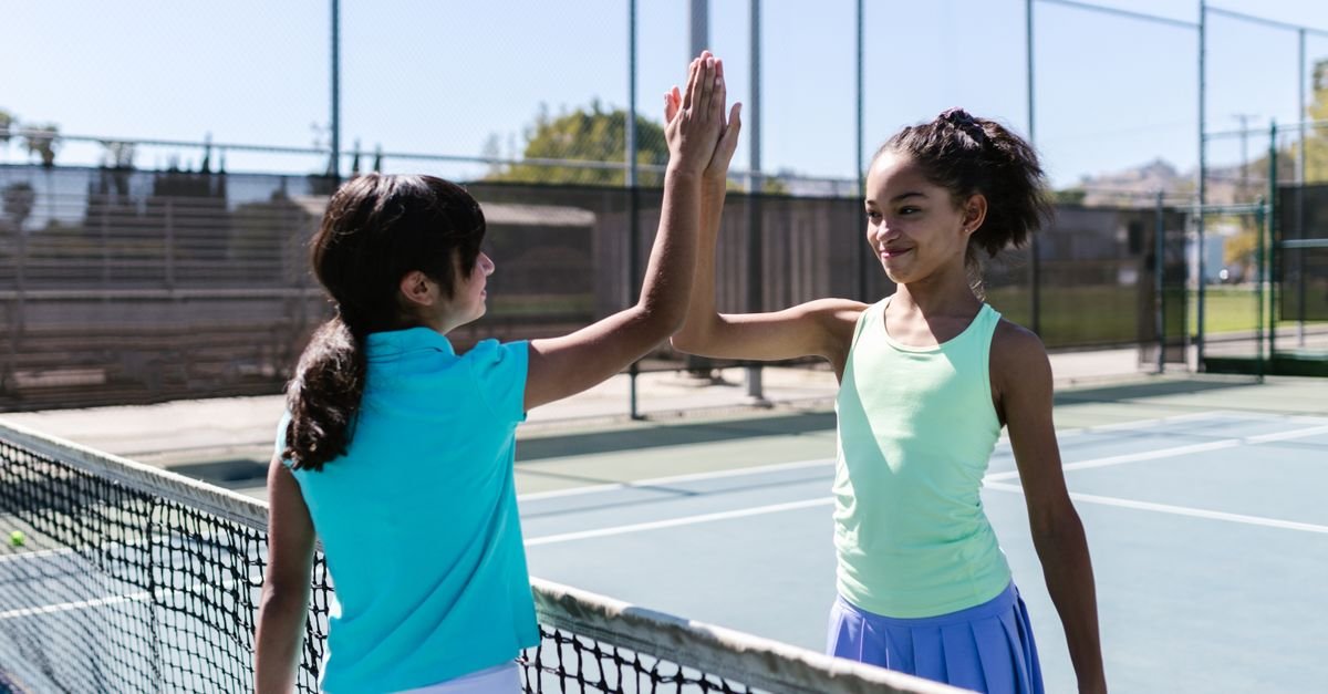 Two teen girls celebrating with a high five on a sunny tennis court.