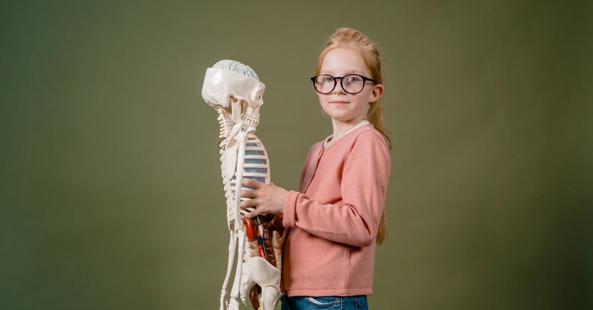 A girl in glasses holding a human skeleton model against a plain background, indoors.