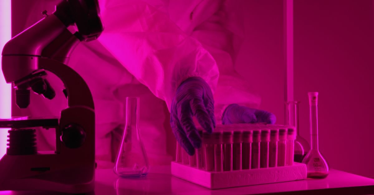 Scientist in a lab coat handling test tubes under pink lighting, using a microscope.