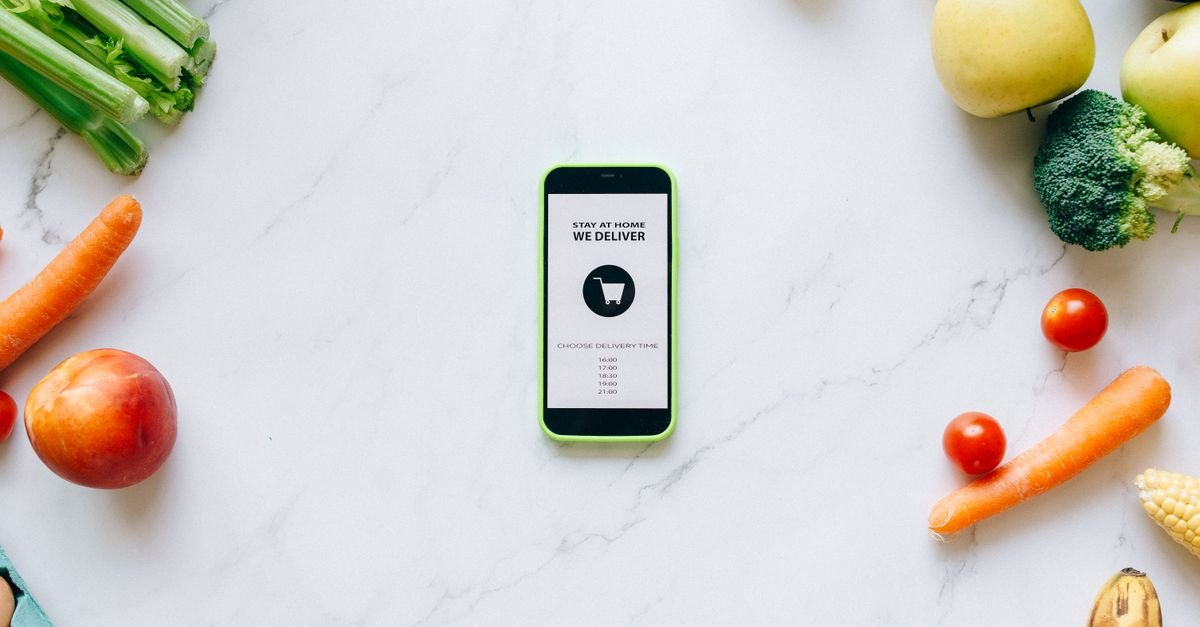 Mobile delivery app surrounded by fresh fruits and vegetables on a marble countertop.