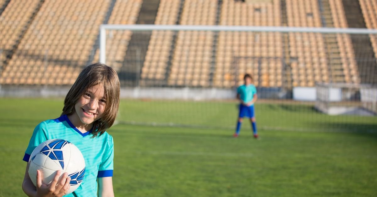 Two kids enjoying a game of soccer at an outdoor field with a stadium backdrop.