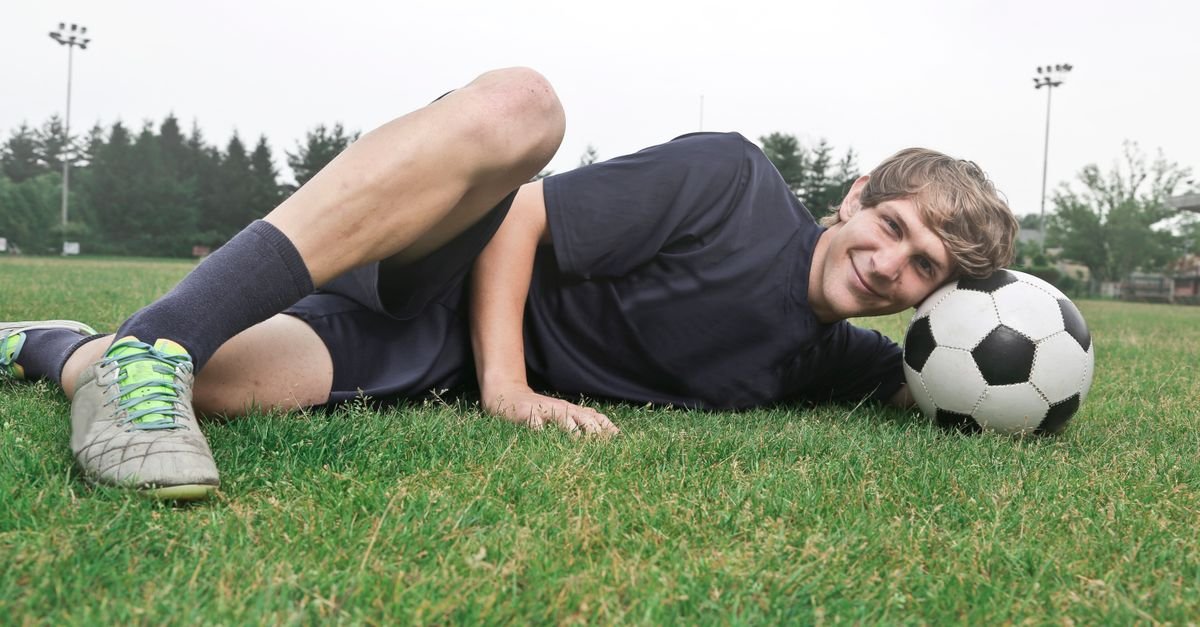 Teen soccer player smiling on grass field resting with a soccer ball.