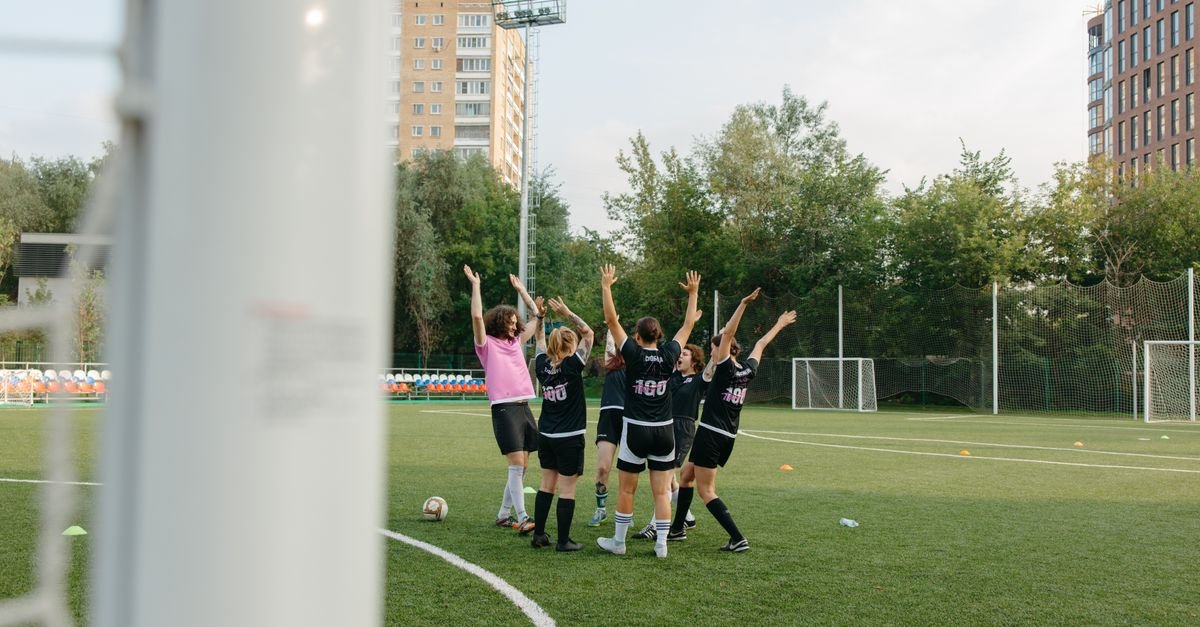 A women's soccer team in black uniforms celebrates a victory on an outdoor soccer field.