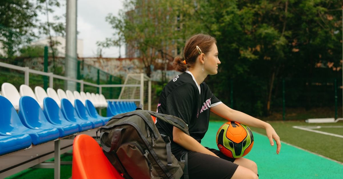 A female soccer player in black uniform sitting on a bench with a ball on a sunny day.
